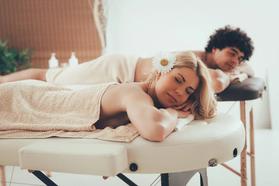 Um casal relaxa em mesas de massagem, cobertas com toalhas; a mulher tem uma flor no cabelo - um cenário ideal para uma massagem para casal ou até mesmo uma Massagem Tântrica.